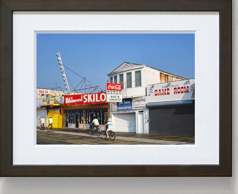 May include: A vintage photograph of a boardwalk scene with a red and yellow sign that reads "Wildwood Skillo". There are four people riding bicycles in front of the sign. The sign is in front of a building with a Coca-Cola sign and a sign that reads "Game Room".