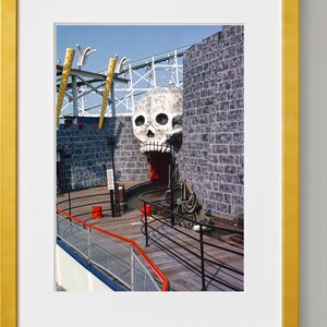Puede incluir: Una gran escultura de calavera blanca con la boca abierta y dientes rojos est&aacute; montada en una pared de ladrillo. La calavera es parte de una atracci&oacute;n de paseo oscuro en un parque de atracciones. La entrada del paseo es visible en el fondo.