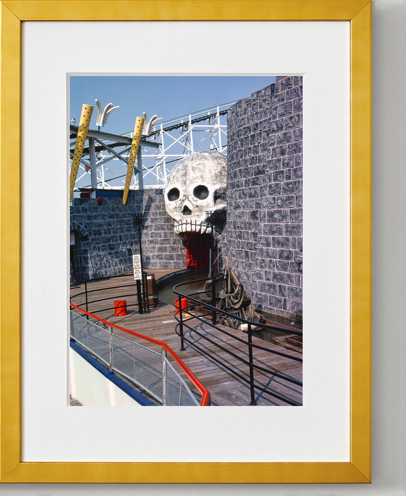 Puede incluir: Una gran escultura de calavera blanca con la boca abierta y dientes rojos est&aacute; montada en una pared de ladrillo. La calavera es parte de una atracci&oacute;n de paseo oscuro en un parque de atracciones. La entrada del paseo es visible en el fondo.
