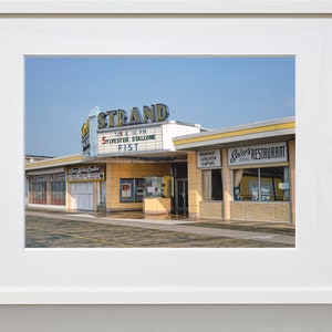 K&ouml;nnte beinhalten: Ein Vintage-Foto eines Kinos namens "The Strand" mit einem Leuchtkasten, der den Film "Fist" mit Sylvester Stallone bewirbt. Das Kino befindet sich neben einem Restaurant namens "Spire's Restaurant".