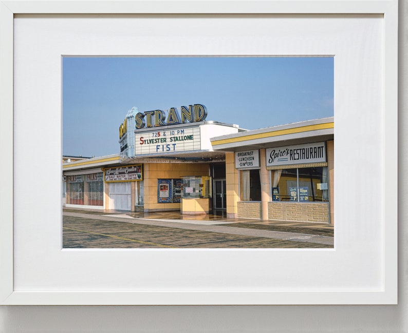 K&ouml;nnte beinhalten: Ein Vintage-Foto eines Kinos namens "The Strand" mit einem Leuchtkasten, der den Film "Fist" mit Sylvester Stallone bewirbt. Das Kino befindet sich neben einem Restaurant namens "Spire's Restaurant".