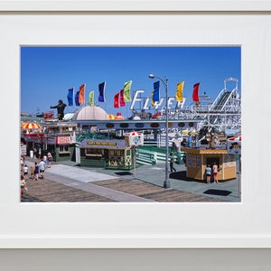 Puede incluir: Una fotograf&iacute;a vintage de un parque de atracciones en un paseo mar&iacute;timo con una monta&ntilde;a rusa, un gran letrero que dice "Fly'er" y varias banderas de colores. Hay gente caminando por el paseo mar&iacute;timo y varias tiendas peque&ntilde;as y puestos de comida.