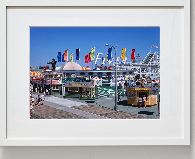 Puede incluir: Una fotograf&iacute;a vintage de un parque de atracciones en un paseo mar&iacute;timo con una monta&ntilde;a rusa, un gran letrero que dice "Fly'er" y varias banderas de colores. Hay gente caminando por el paseo mar&iacute;timo y varias tiendas peque&ntilde;as y puestos de comida.
