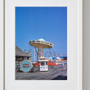 May include: A vintage photograph of a fairground ride on a pier. The ride is a carousel with a blue and white canopy and swings. The ride is in the foreground of the image, with a blue sky in the background. There is a sign that says "Down Under Cart Track" in the foreground.