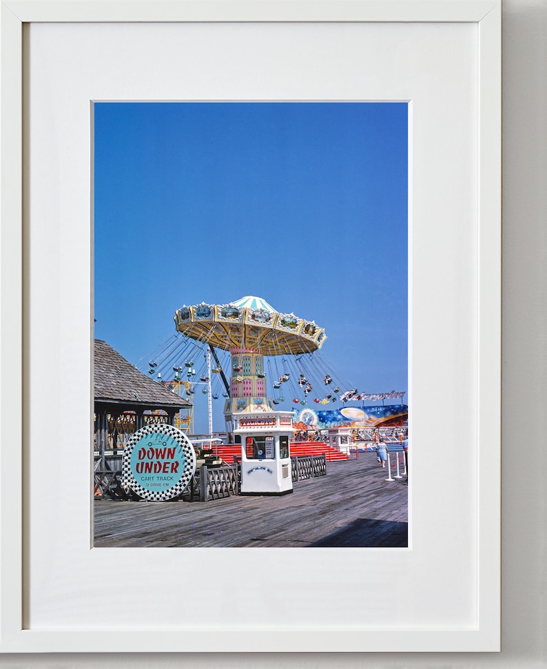 May include: A vintage photograph of a fairground ride on a pier. The ride is a carousel with a blue and white canopy and swings. The ride is in the foreground of the image, with a blue sky in the background. There is a sign that says "Down Under Cart Track" in the foreground.