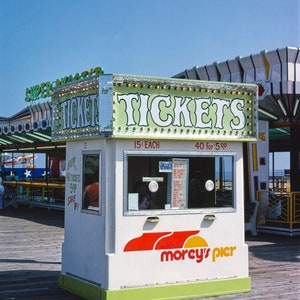 May include: A white ticket booth with green and red accents, displaying the word "TICKETS" in large, illuminated letters. The booth has a window and the "Morey's Pier" logo. The sky is blue.
