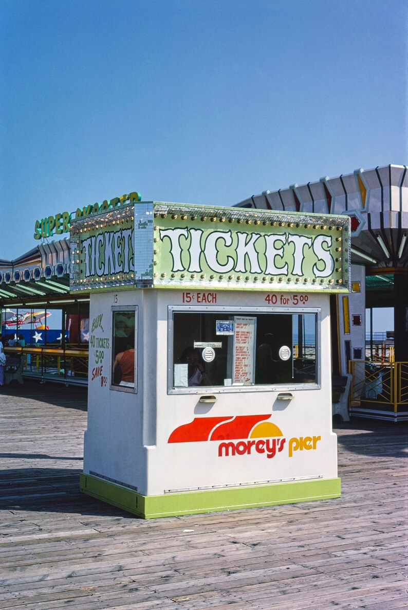 May include: A white ticket booth with green and red accents, displaying the word "TICKETS" in large, illuminated letters. The booth has a window and the "Morey's Pier" logo. The sky is blue.