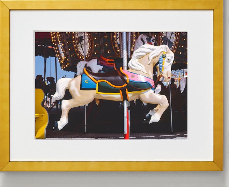 May include: A white carousel horse with a brown saddle, pink and green trim, and a blue and yellow mane. The horse is on a carousel with lights and other horses in the background.