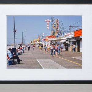 Peut inclure: Une photographie vintage d'une promenade avec des boutiques et des enseignes color&eacute;es. Les boutiques portent des noms comme "Grandpa's", "Fun Shop" et "Snow White". Des gens se prom&egrave;nent sur la promenade, certains sont assis sur des bancs.