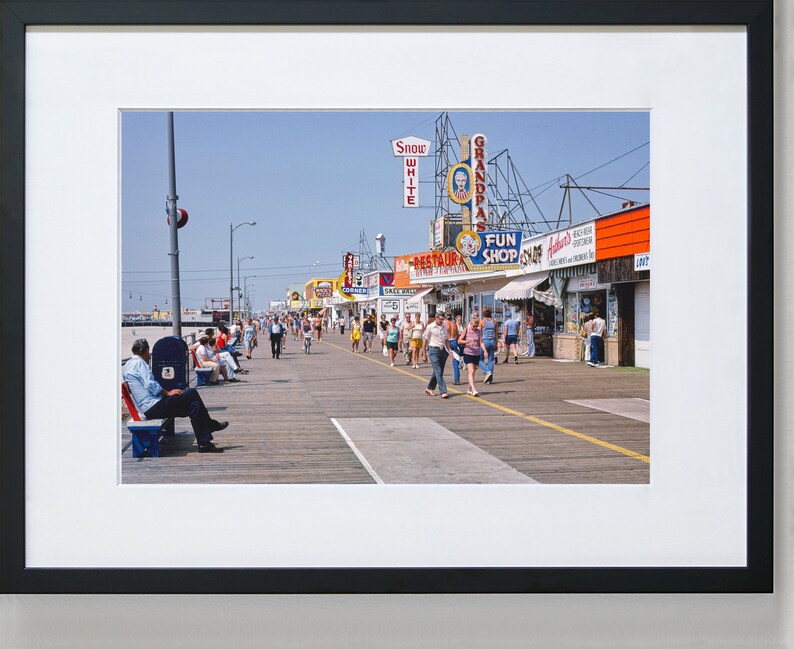 Peut inclure: Une photographie vintage d'une promenade avec des boutiques et des enseignes color&eacute;es. Les boutiques portent des noms comme "Grandpa's", "Fun Shop" et "Snow White". Des gens se prom&egrave;nent sur la promenade, certains sont assis sur des bancs.