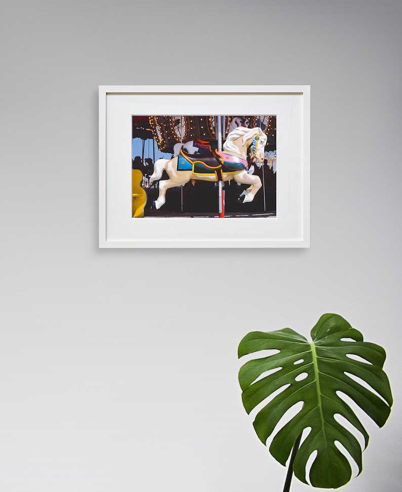 May include: A white carousel horse with a colorful saddle and harness. The horse is in motion, with its head turned to the side and its tail flowing behind it. The horse is part of a carousel, and there are other horses and lights in the background.