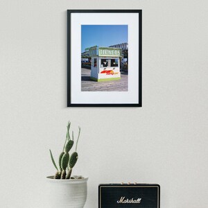 May include: Framed photograph of a ticket booth with the word "TICKETS" in green lettering. The booth has a white and green exterior with the words "morey's pier" in red and yellow. A cactus in a white pot and a black Marshall speaker are in the foreground.