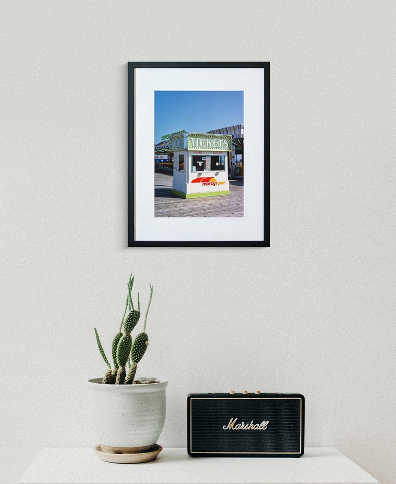 May include: Framed photograph of a ticket booth with the word "TICKETS" in green lettering. The booth has a white and green exterior with the words "morey's pier" in red and yellow. A cactus in a white pot and a black Marshall speaker are in the foreground.
