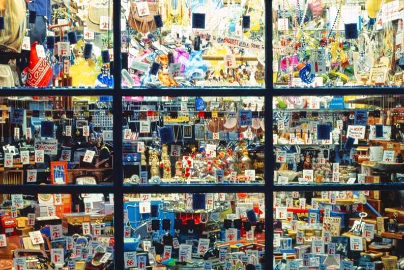 May include: A shop window display packed with a variety of items for sale, including jewellery, trinkets, and small toys. The window is divided into sections, each displaying a different selection of goods. Numerous price tags are visible, indicating the items are available to buy.