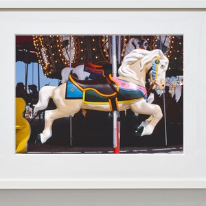 May include: A white carousel horse with a brown saddle, colorful trim, and a blue and gold eye. The horse is on a carousel with lights and other horses in the background.