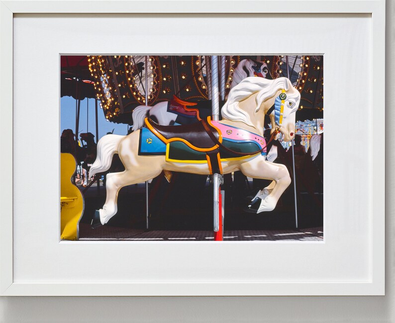 May include: A white carousel horse with a brown saddle, colorful trim, and a blue and gold eye. The horse is on a carousel with lights and other horses in the background.