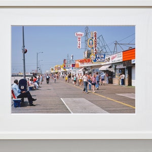 Peut inclure: Une photographie vintage d'une promenade avec des boutiques et des enseignes color&eacute;es. Des gens se prom&egrave;nent sur la promenade, certains sont assis sur des bancs. Les enseignes comprennent "Snow White", "Grandpa's", "Fun Shop" et "Restaurant".
