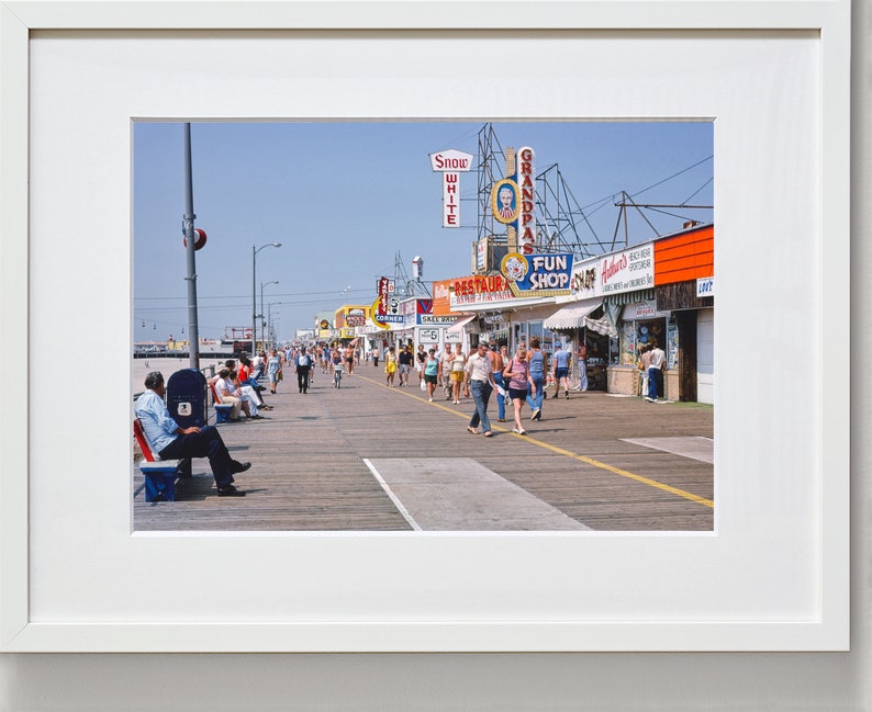 Peut inclure: Une photographie vintage d'une promenade avec des boutiques et des enseignes color&eacute;es. Des gens se prom&egrave;nent sur la promenade, certains sont assis sur des bancs. Les enseignes comprennent "Snow White", "Grandpa's", "Fun Shop" et "Restaurant".