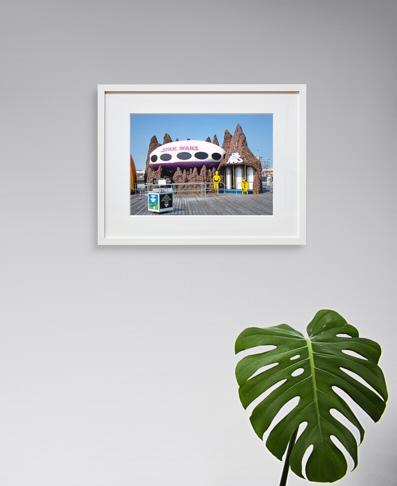 May include: A framed photograph of a Star Wars themed amusement park ride. The ride is a large, white, flying saucer with purple accents. The saucer is hovering over a brown, rocky landscape. The words "Star Wars" are printed in pink on the saucer.