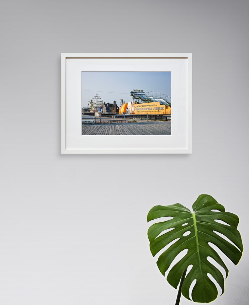 May include: A framed photograph of a boardwalk with a large sign that reads "Mickey's Kiddie Park Presents...  The Old Country Jubilee". The sign is yellow and orange with black lettering. The boardwalk is made of wood and there are several buildings in the background.