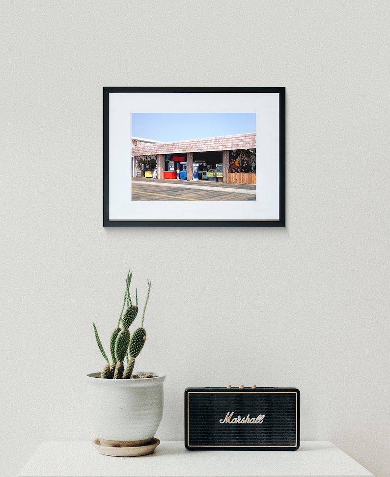 Pode incluir: Uma fotografia vintage de uma pequena loja com uma marquise vermelha e v&aacute;rios jogos de arcade no interior. A loja est&aacute; localizada em um estacionamento pavimentado.