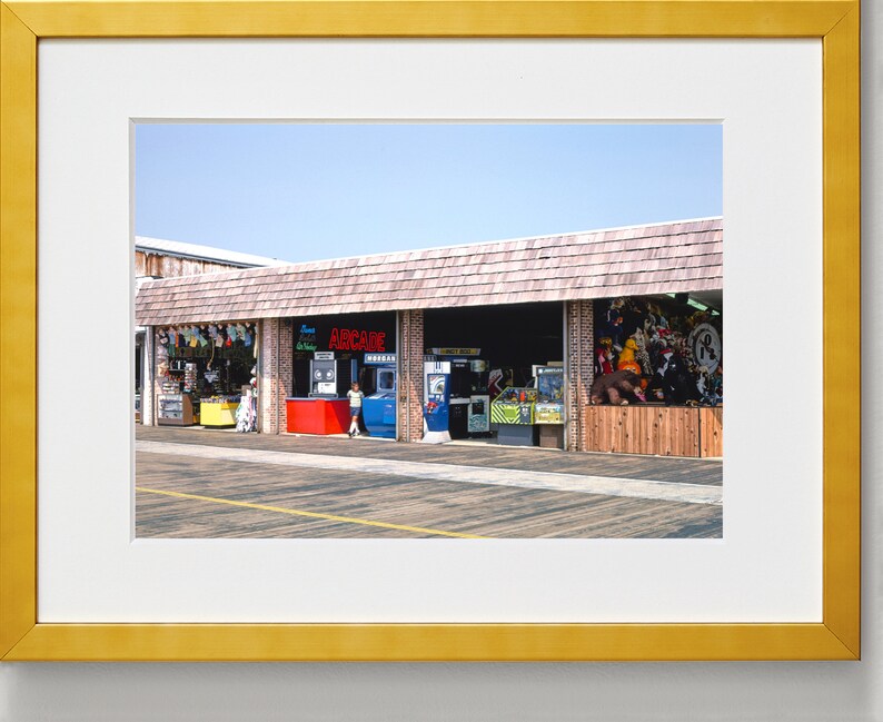 Pode incluir: Uma loja de arcade bem iluminada com uma placa vermelha que diz "ARCADE" e uma variedade de jogos de arcade vis&iacute;veis atrav&eacute;s das janelas. A loja &eacute; feita de madeira e tijolo e tem um toldo de madeira.