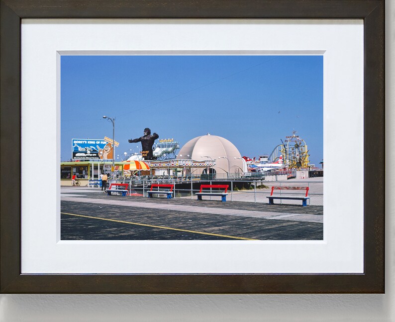 Op de afbeelding: Een vintage foto van een pretpark aan een boulevard met een groot gorillabeeld, een wit koepelvormig gebouw en een achtbaan op de achtergrond. De foto is gemaakt op een zonnige dag met een blauwe lucht.