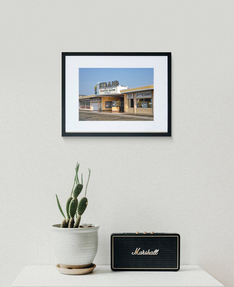K&ouml;nnte beinhalten: Ein Vintage-Foto eines gelben Geb&auml;udes mit einem Schild, auf dem "The Strand Theatre" in einer Retro-Schrift steht. Das Geb&auml;ude hat ein gro&szlig;es Vordach mit den Worten "Coming Soon" und "The Tall Texan" in roten Buchstaben. Das Foto ist in einem schwarzen Rahmen gerahmt.