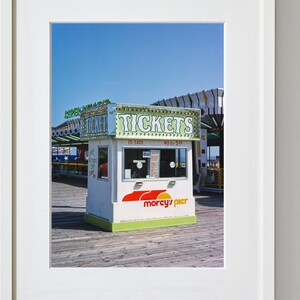 May include: A photograph of a ticket booth on a pier. The booth is white with green and red accents, and the word "TICKETS" is prominently displayed. The booth also has the text "morcy's pier". The sky is blue.