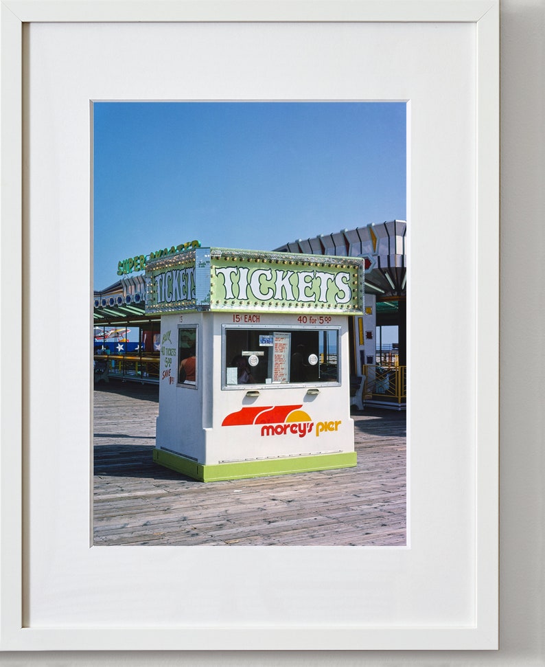 May include: A photograph of a ticket booth on a pier. The booth is white with green and red accents, and the word "TICKETS" is prominently displayed. The booth also has the text "morcy's pier". The sky is blue.