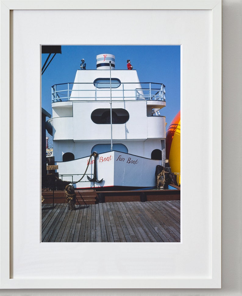 May include: A white boat with a red and blue striped roof and the words "Fun Boat" written on the side. The boat is docked at a wooden pier.