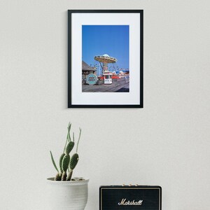 May include: A framed photograph of a vintage amusement park ride with a blue sky in the background. The ride is a carousel with a white canopy and colourful seats. The ride is located on a boardwalk with a wooden floor and a few buildings in the background.