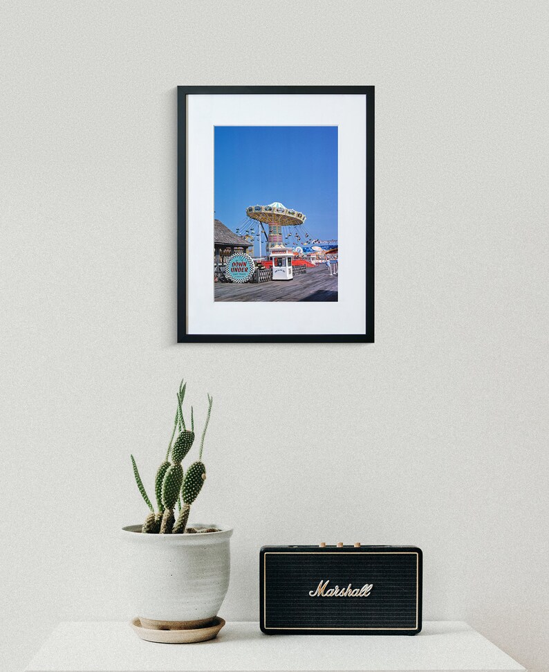 May include: A framed photograph of a vintage amusement park ride with a blue sky in the background. The ride is a carousel with a white canopy and colourful seats. The ride is located on a boardwalk with a wooden floor and a few buildings in the background.
