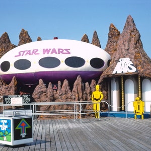 May include: A Star Wars themed amusement park ride with a white and purple spaceship, brown rock formations, and yellow figures of C-3PO and R2-D2. The sign reads "Star Wars".