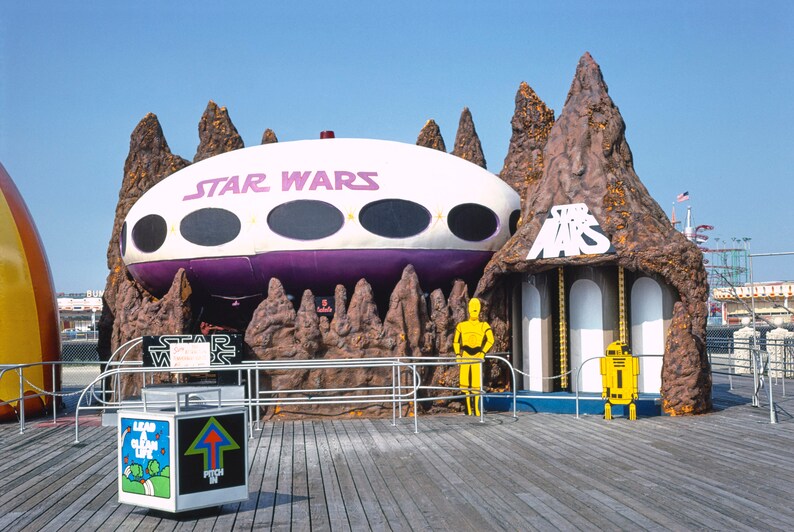 May include: A Star Wars themed amusement park ride with a white and purple spaceship, brown rock formations, and yellow figures of C-3PO and R2-D2. The sign reads "Star Wars".