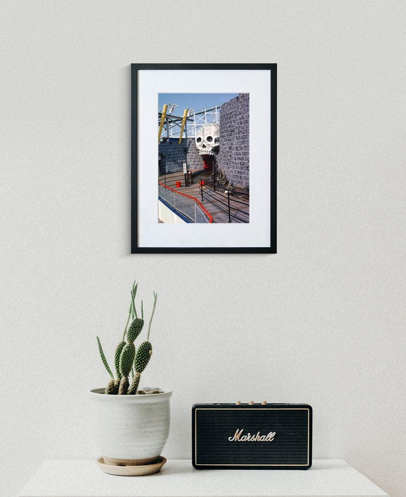 Puede incluir: Una fotograf&iacute;a enmarcada de una gran escultura de calavera blanca con la boca abierta, sobre un fondo de pared de ladrillo gris. La calavera forma parte de una estructura m&aacute;s grande, posiblemente una atracci&oacute;n de un parque de atracciones, con una pasarela de madera que conduce a ella.
