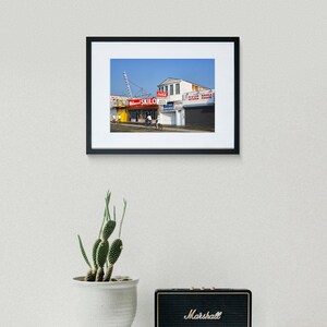 May include: A framed photograph of a colorful building with a red sign that reads "Skilo's" and a blue sign that reads "Game Room". The building is located on a street with a blue sky in the background.