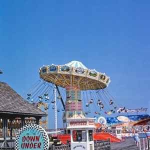 May include: A vintage photograph of a carnival amusement park with a colourful carousel ride in the centre. The ride is called "Down Under" and is a cart track u-drive-em. There is a white ticket booth with the word "Tickets" on it.
