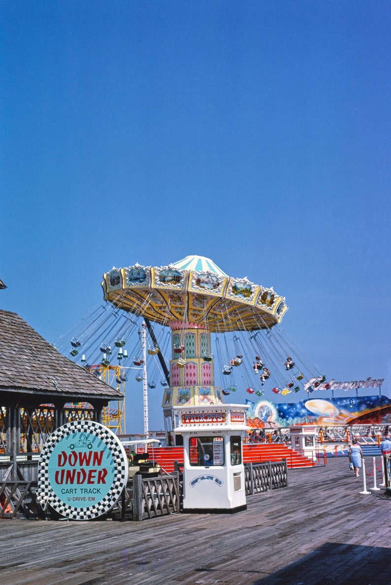 May include: A vintage photograph of a carnival amusement park with a colourful carousel ride in the centre. The ride is called "Down Under" and is a cart track u-drive-em. There is a white ticket booth with the word "Tickets" on it.