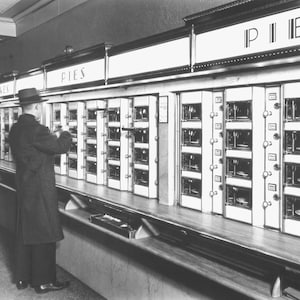 May include: A vintage black and white photo of a person standing in front of a row of vending machines. The machines are labeled "PIES" and have glass doors with rows of pies inside.