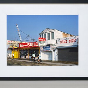 May include: A vintage photo of a boardwalk storefront with a red and yellow sign that reads "Skilo" and a Coca-Cola sign. There are four people riding bicycles in front of the store.