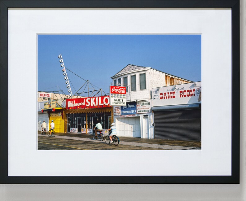 May include: A vintage photo of a boardwalk storefront with a red and yellow sign that reads "Skilo" and a Coca-Cola sign. There are four people riding bicycles in front of the store.