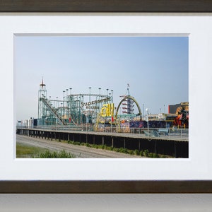 Op de afbeelding: Een ingelijste print van een pretpark aan zee met een achtbaan en andere attracties. De achtbaan is groen en zilverkleurig, met de tekst "Supersonic Roller Coaster" zichtbaar. De lucht is helderblauw.