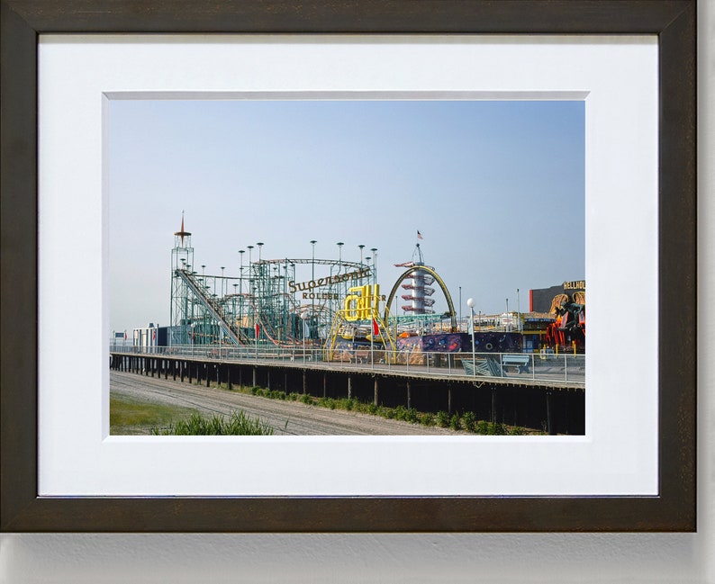 Op de afbeelding: Een ingelijste print van een pretpark aan zee met een achtbaan en andere attracties. De achtbaan is groen en zilverkleurig, met de tekst "Supersonic Roller Coaster" zichtbaar. De lucht is helderblauw.