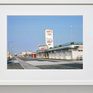 May include: A vintage photograph of a boardwalk with a sign for Hunt's Pier and Skyline Golf. The sign is red and white with a green golf ball. The boardwalk is lined with shops and businesses, including one with a sign that says "Jaws 2".