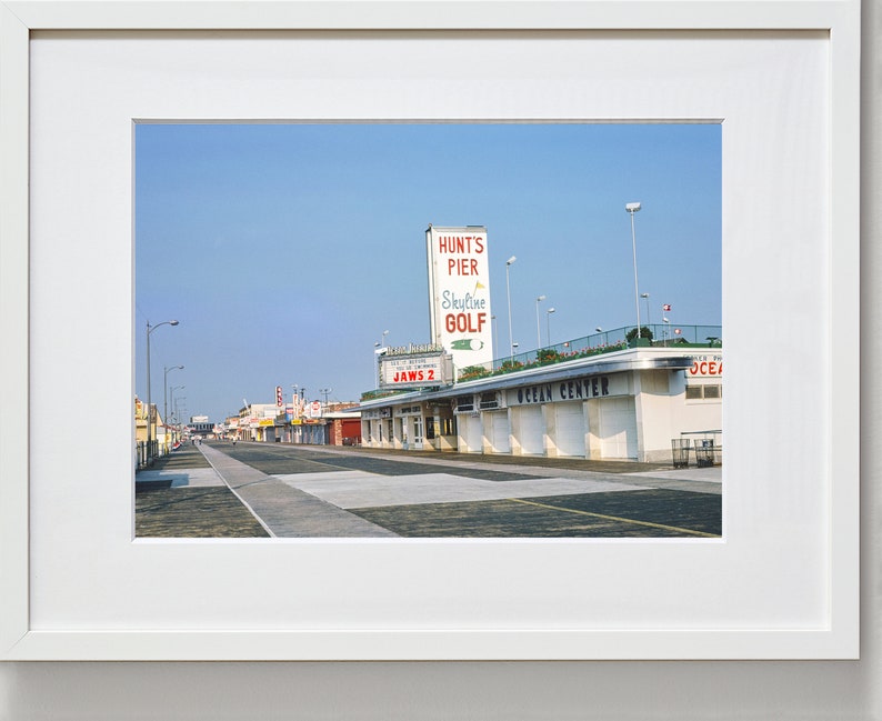 May include: A vintage photograph of a boardwalk with a sign for Hunt's Pier and Skyline Golf. The sign is red and white with a green golf ball. The boardwalk is lined with shops and businesses, including one with a sign that says "Jaws 2".