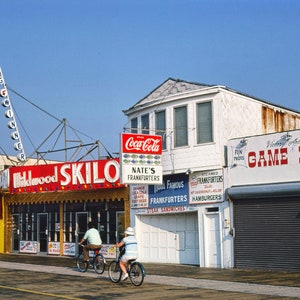 May include: A brightly colored storefront with a red sign that reads "Skilo" and a sign for "Nate's Frankfurters". There is a white building with a sign that reads "Victory Arcade Game Room".  Three people are riding bicycles in front of the storefronts.
