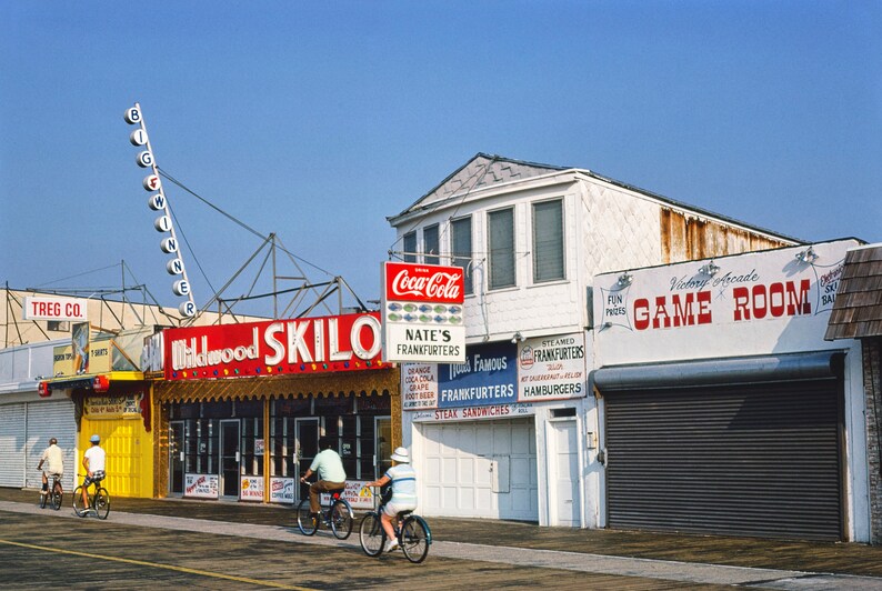 May include: A brightly colored storefront with a red sign that reads "Skilo" and a sign for "Nate's Frankfurters". There is a white building with a sign that reads "Victory Arcade Game Room".  Three people are riding bicycles in front of the storefronts.