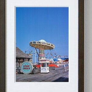 May include: A vintage photograph of a fairground ride on a pier. The ride is a carousel with a blue and white canopy and swings. There is a small white building with a sign that says "Tickets". The sign "Down Under Cart Track" is visible in the foreground.