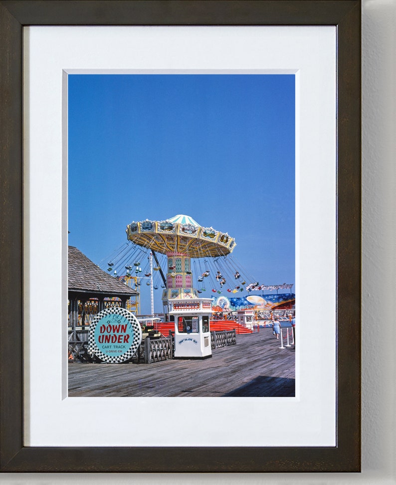 May include: A vintage photograph of a fairground ride on a pier. The ride is a carousel with a blue and white canopy and swings. There is a small white building with a sign that says "Tickets". The sign "Down Under Cart Track" is visible in the foreground.